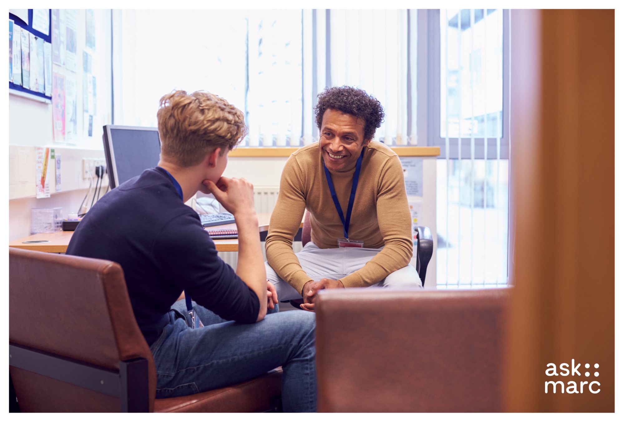a picture of two men sitting in an office talking, One is facing the camera, the other is cacing him away from the camera.